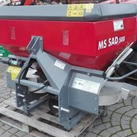 Czerwony rozsiewacz nawozów MS SAD 500 na szarej ramie, widoczny na palecie. Urządzenie z szarym, płaskim daszkiem, przeznaczone do montażu na ciągniku.