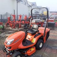 Pomarańczowa kosiarka samojezdna Kubota GR1600 z czarnym siedzeniem i koszem na trawę, na tle innych maszyn rolniczych i budynku w lekko zamglony dzień.