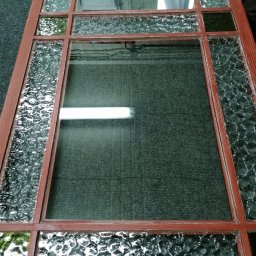 Zakład Szklarski SZKLARZTOM Maciej Tomaszewicz - Fragment drewnianej ramy okiennej z szybami o różnej fakturze, w tym jedna szyba przezroczysta, na szarym tle