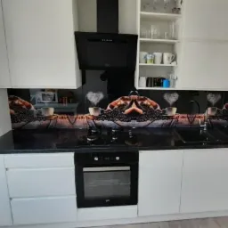 Szklany panel kuchenny z motywem kawy, zamontowany nad blatem i pomiędzy białymi szafkami, z widoczną płytą grzewczą i piekarnikiem.