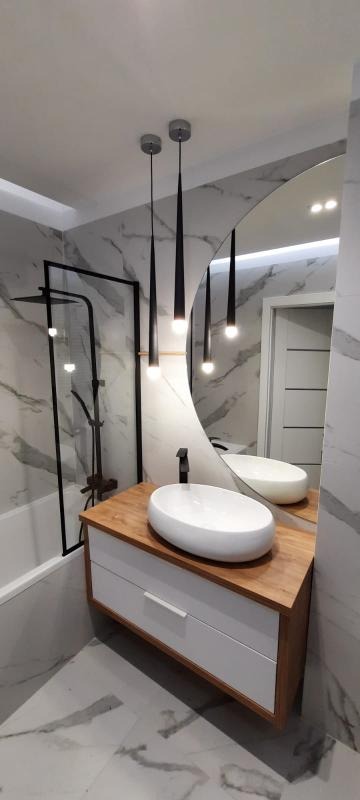 Nowoczesna łazienka z owalną umywalką nablatową, drewnianym blatem, białymi szafkami, okrągłym lustrem z podświetleniem LED, czarną armaturą i kabiną prysznicową w czarnej ramie na tle marmurowych...