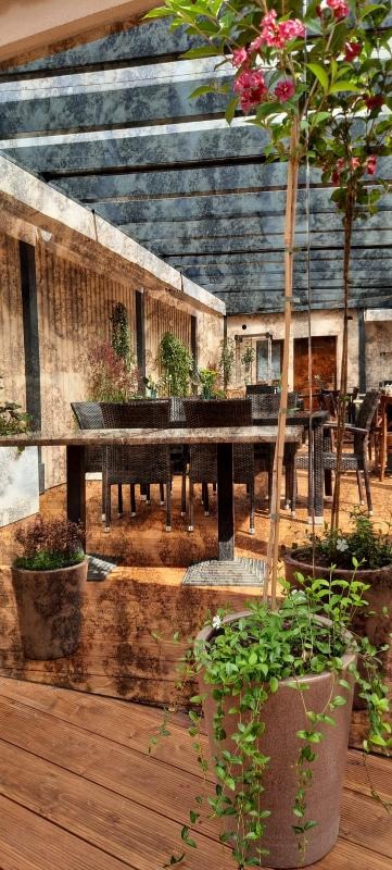 Zewnętrzna restauracja z drewnianym tarasem, stołami i krzesłami z rattanu, rośliny w donicach i zadaszenie z metalowych belek.