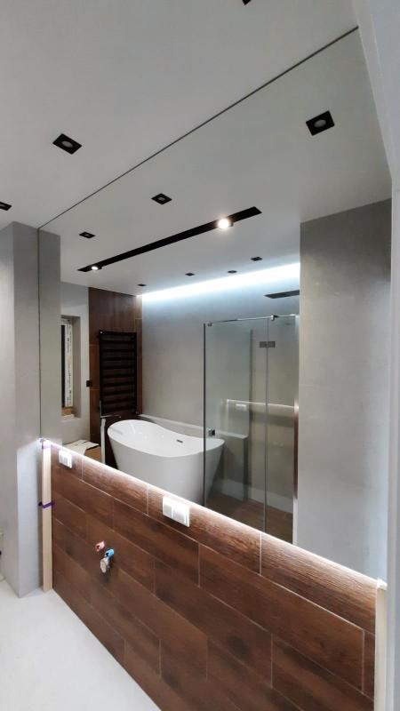 Nowoczesna łazienka z owalną wanną, szklaną kabiną prysznicową i drewnopodobną zabudową ściany z oświetleniem LED wzdłuż krawędzi, widok zza niskiej ścianki działowej.