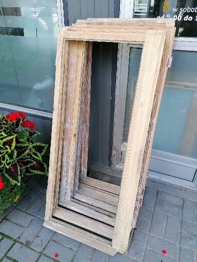 Stos drewnianych ram okiennych, widoczne ślady zużycia i odbarwienia, ustawione na brukowanym chodniku, obok doniczka z czerwono-zielonymi kwiatami, w tle okno wystawowe.