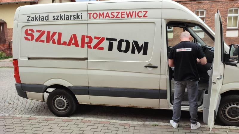 Biały samochód dostawczy z czerwono-czarnym napisem 'Zakład szklarski TOMASZEWICZ SZKLARZTOM', mężczyzna w czarnej koszulce z napisem 'SZKLARZTOM' wchodzi do pojazdu.