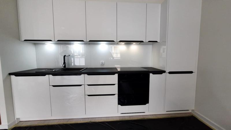 Nowoczesna, biała kuchnia z czarnym blatem i szklanym panelem ściennym, oświetlona punktowymi lampami podszafkowymi, z widoczną czarną baterią i płytą indukcyjną.