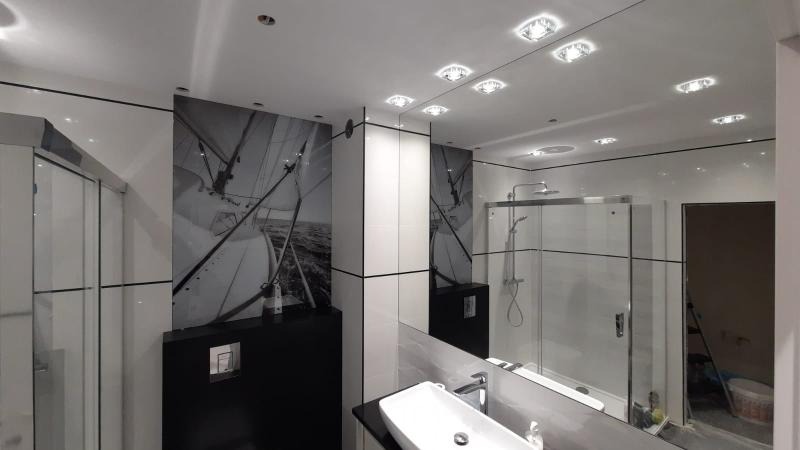 Nowoczesna łazienka z dużym lustrem na ścianie, odbijającym kabinę prysznicową, oraz fototapetą z motywem żaglówki, biała umywalka na czarnym blacie.