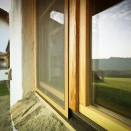 Drewniana rama okienna z zamontowaną siatką przeciw owadom, widok na wiejski krajobraz odbity w szybie okna