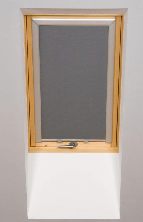 Szara roleta zwijana w drewnianej ramie okna dachowego, widok z bliska na mechanizm blokujący.