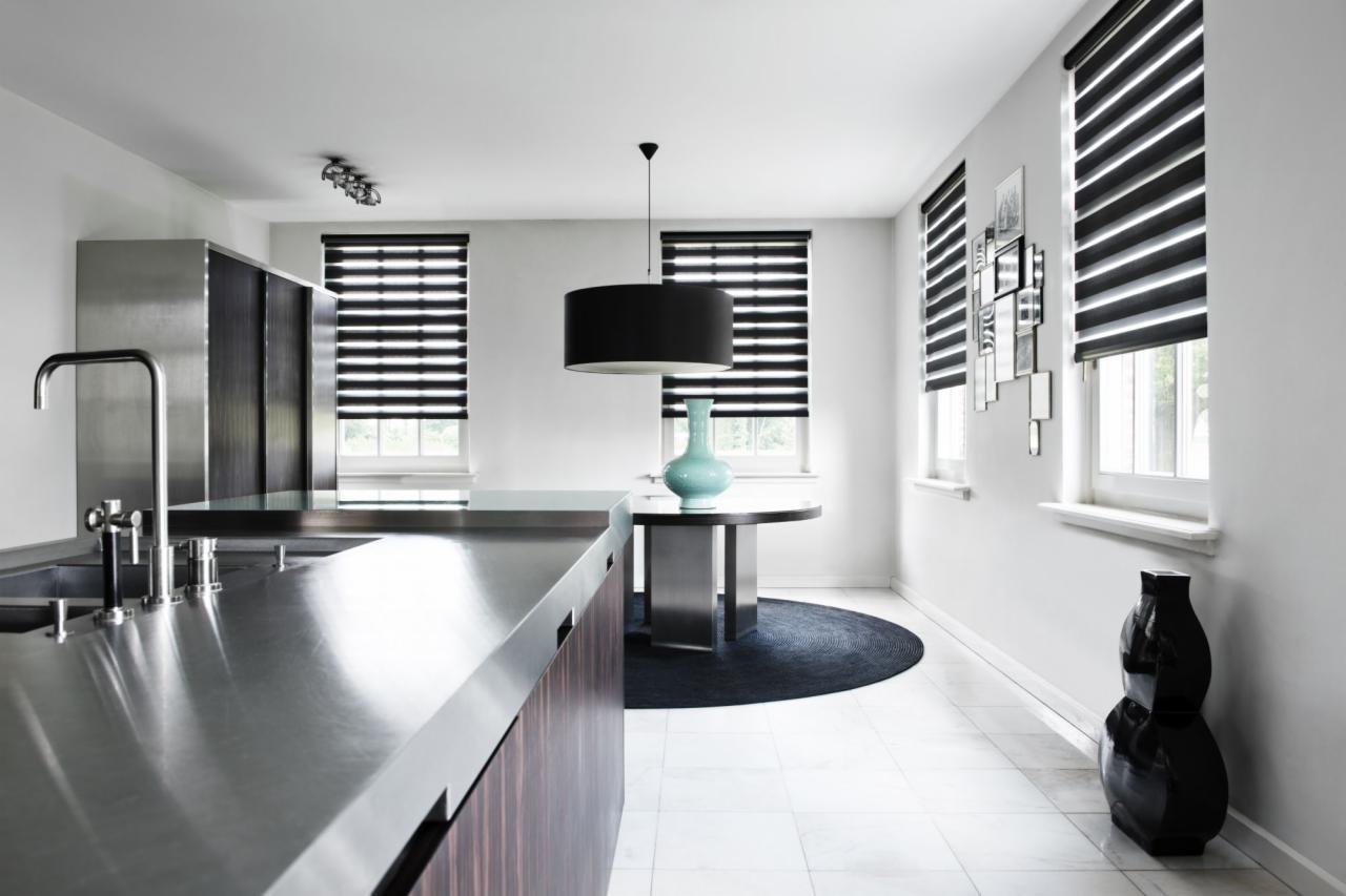 Nowoczesna kuchnia z metalowym blatem i oknami przesłoniętymi roletami w czarno-białe pasy, okrągły stół z turkusowym wazonem i czarnym dywanem.