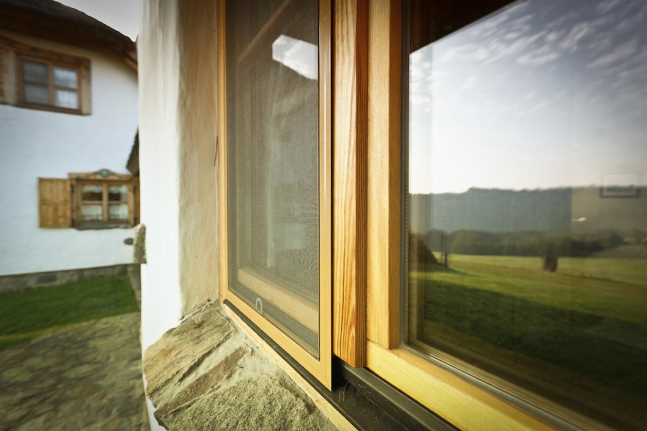 Drewniana rama okienna z zamontowaną siatką przeciw owadom, widok na wiejski krajobraz odbity w szybie okna