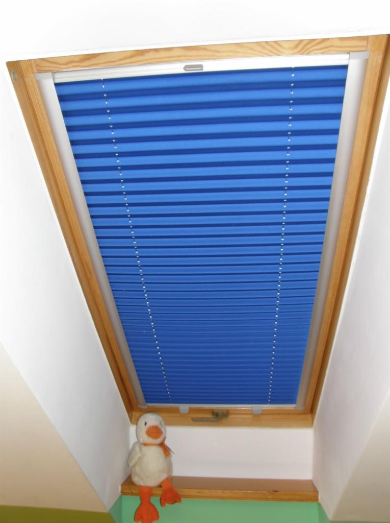Niebieska plisa okienna w drewnianej ramie okna dachowego, z pluszową kaczką siedzącą na parapecie.