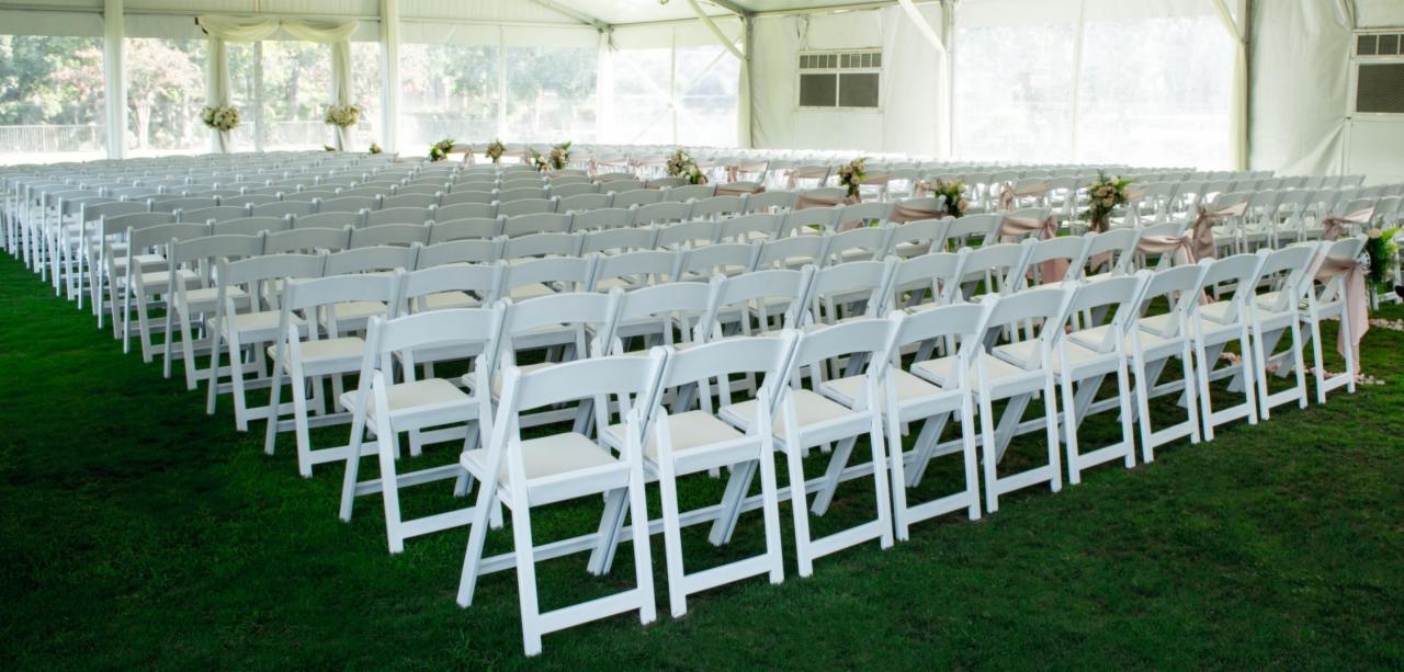 Ustawione rzędy białych krzeseł z poduszkami wewnątrz namiotu, udekorowane kwiatami i wstążkami, przygotowane na ceremonię.