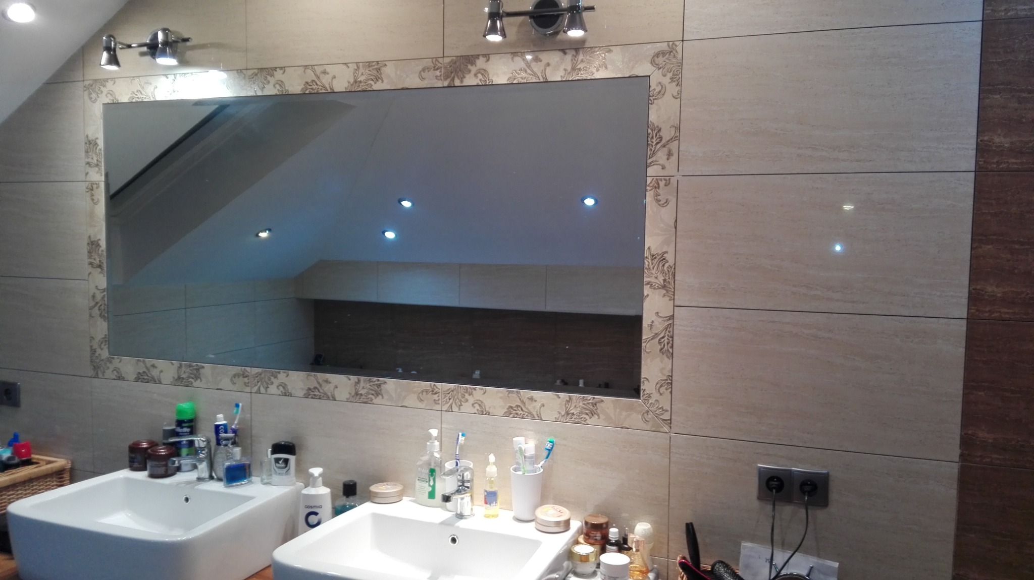 Dwie umywalki w łazience z beżowymi płytkami i dużym lustrem z ozdobną ramą, oświetlone lampami sufitowymi.