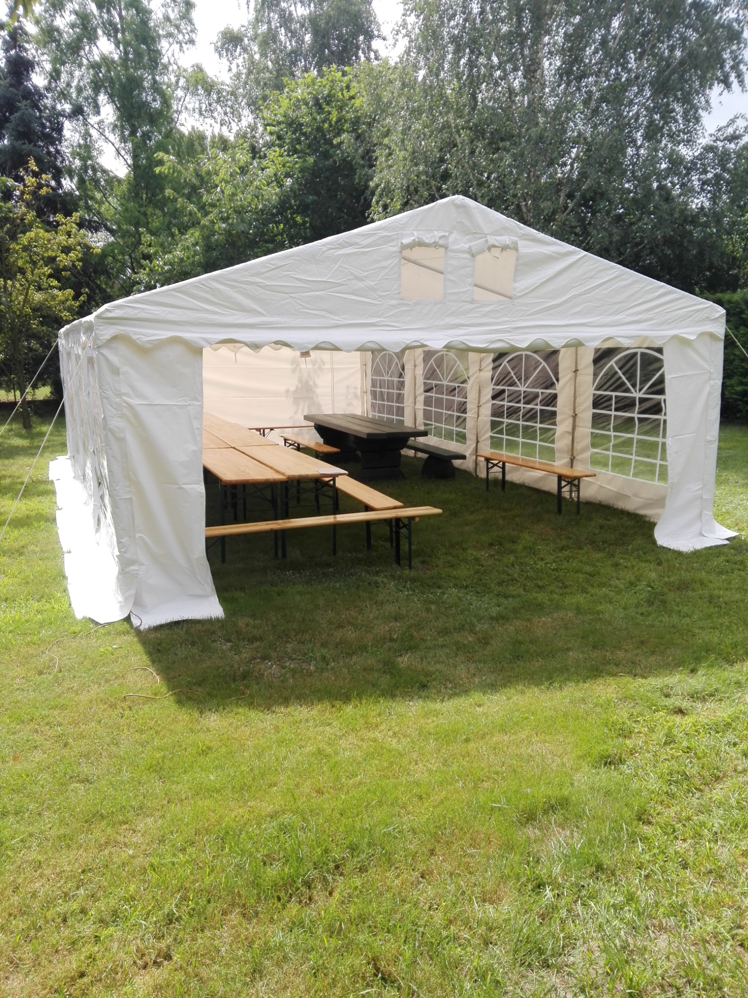 Biały namiot imprezowy z drewnianymi stołami i ławkami wewnątrz, rozstawiony na trawniku, widok z zewnątrz w słoneczny dzień.