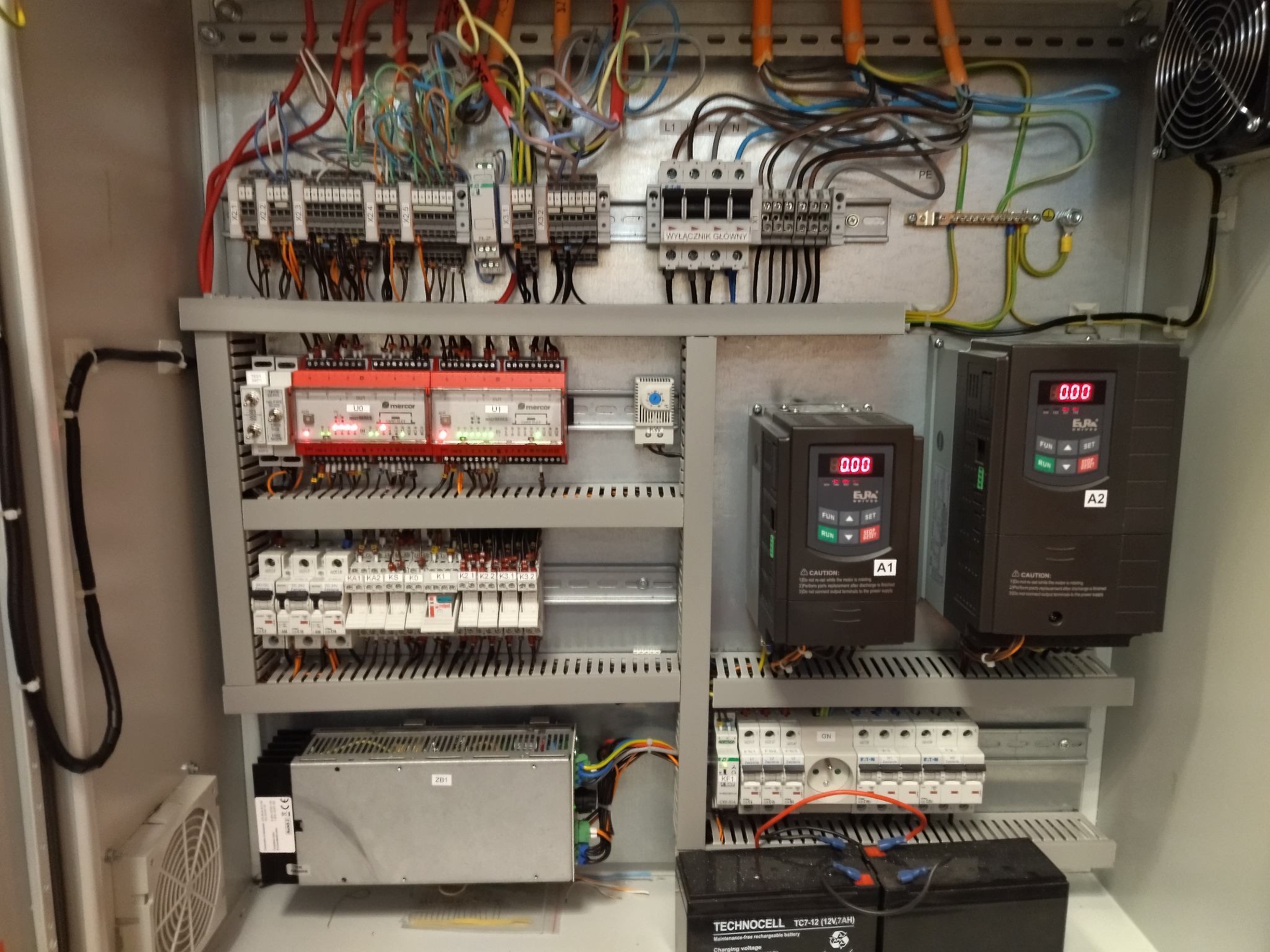 Wnętrze szafy sterowniczej z widocznymi elementami elektrycznymi, w tym sterownikami, bezpiecznikami, przewodami i baterią akumulatorową na dole.