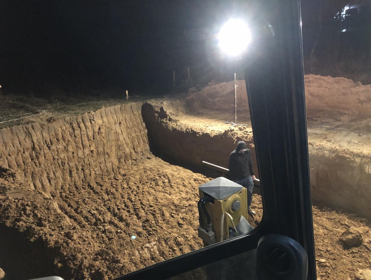 Głęboki wykop w ziemi widziany z kabiny maszyny budowlanej, oświetlony mocnym światłem reflektora, z pracownikiem w tle, wykonany nocą.