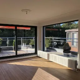 Nowoczesne, przesuwne okna z czarnymi ramami otwierają się na taras z roślinami w donicach, widok na zieleń z wnętrza z parkietem i grzejnikiem.