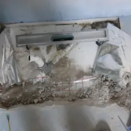 Montaż odpływu liniowego w trakcie remontu łazienki, widoczne rury, folia w płynie i buty robocze na tle surowego betonu.