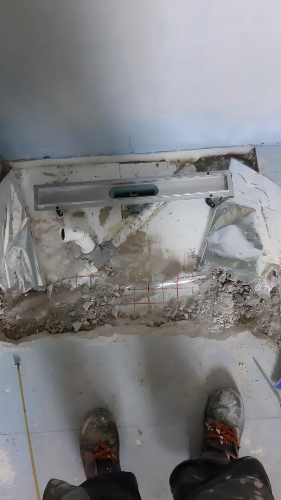 Montaż odpływu liniowego w trakcie remontu łazienki, widoczne rury, folia w płynie i buty robocze na tle surowego betonu.