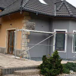 Dom w Łodygowicach częściowo otynkowany na szaro, z widocznym rusztowaniem i fragmentem elewacji w kolorze beżowym, prezentujący etap prac wykończeniowych elewacji.