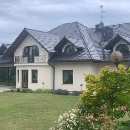 Jacek Michalik - Elegancki dom jednorodzinny z jasną elewacją, ciemnym dachem z lukarnami i balkonem z kutą balustradą. Przed domem zadbany ogród z krzewami i kwiatami.