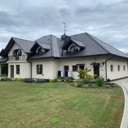 Jacek Michalik - Nowoczesny dom jednorodzinny z jasną elewacją i ciemnym dachem z lukarnami o półokrągłych oknach. W ogrodzie ścieżka z kostki, rabaty i dwie jednostki pomp ciepła.