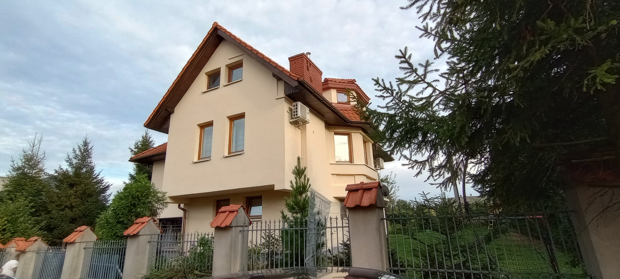 Beżowy dom jednorodzinny z brązowym dachem, widoczny zza metalowego ogrodzenia, z zainstalowanymi klimatyzatorami na elewacji.