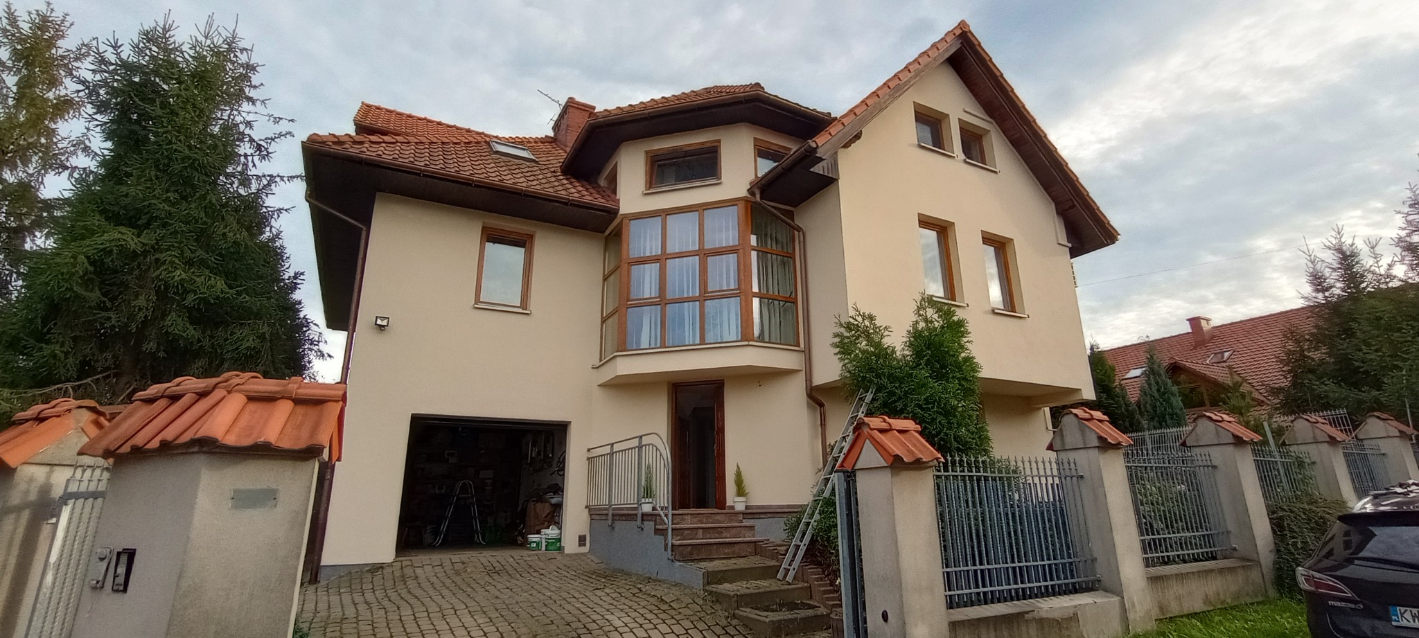 Beżowy dom jednorodzinny z brązową stolarką okienną i czerwoną dachówką, widok z zewnątrz po renowacji, z widoczną drabiną opartą o ogrodzenie i otwartym garażem z narzędziami.