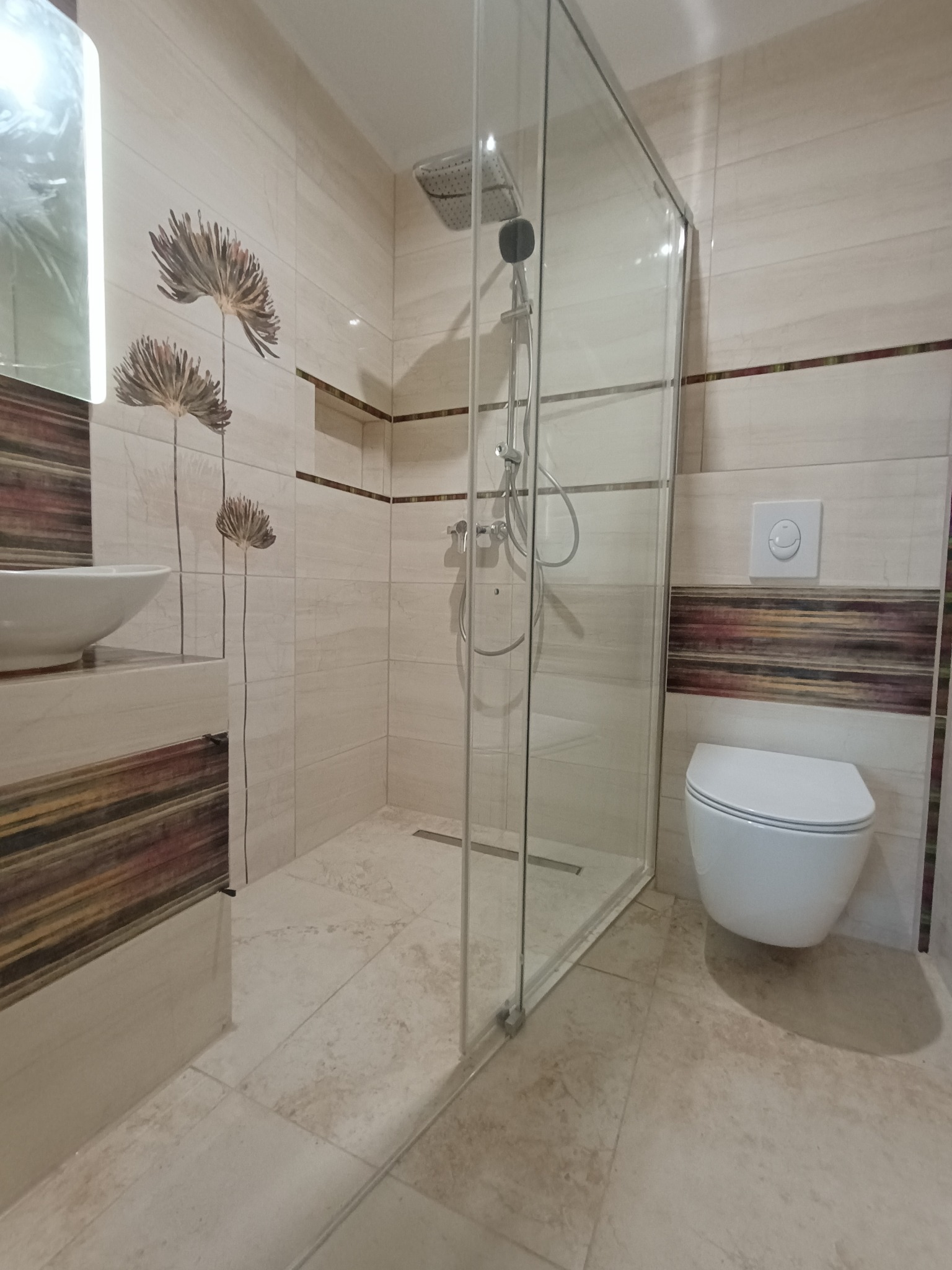 Wykończona łazienka z kabiną prysznicową typu walk-in, umywalką nablatową i podwieszaną toaletą. Beżowe płytki z dekoracyjnym motywem kwiatowym i poziomymi pasami.