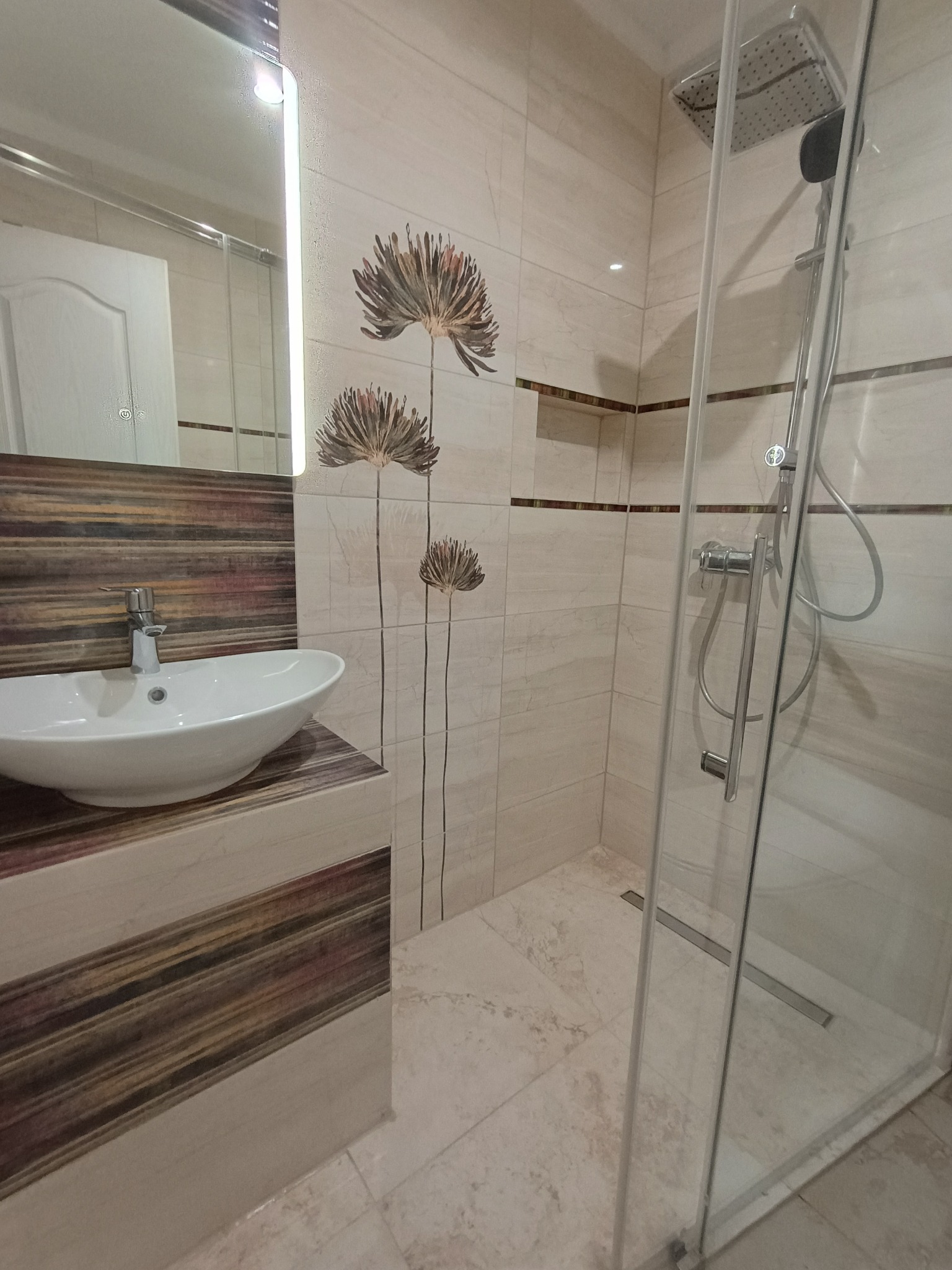 Nowoczesna łazienka z kabiną prysznicową typu walk-in, umywalką nablatową i lustrem z podświetleniem LED. Beżowe płytki z motywem kwiatowym i drewnopodobne wykończenia.