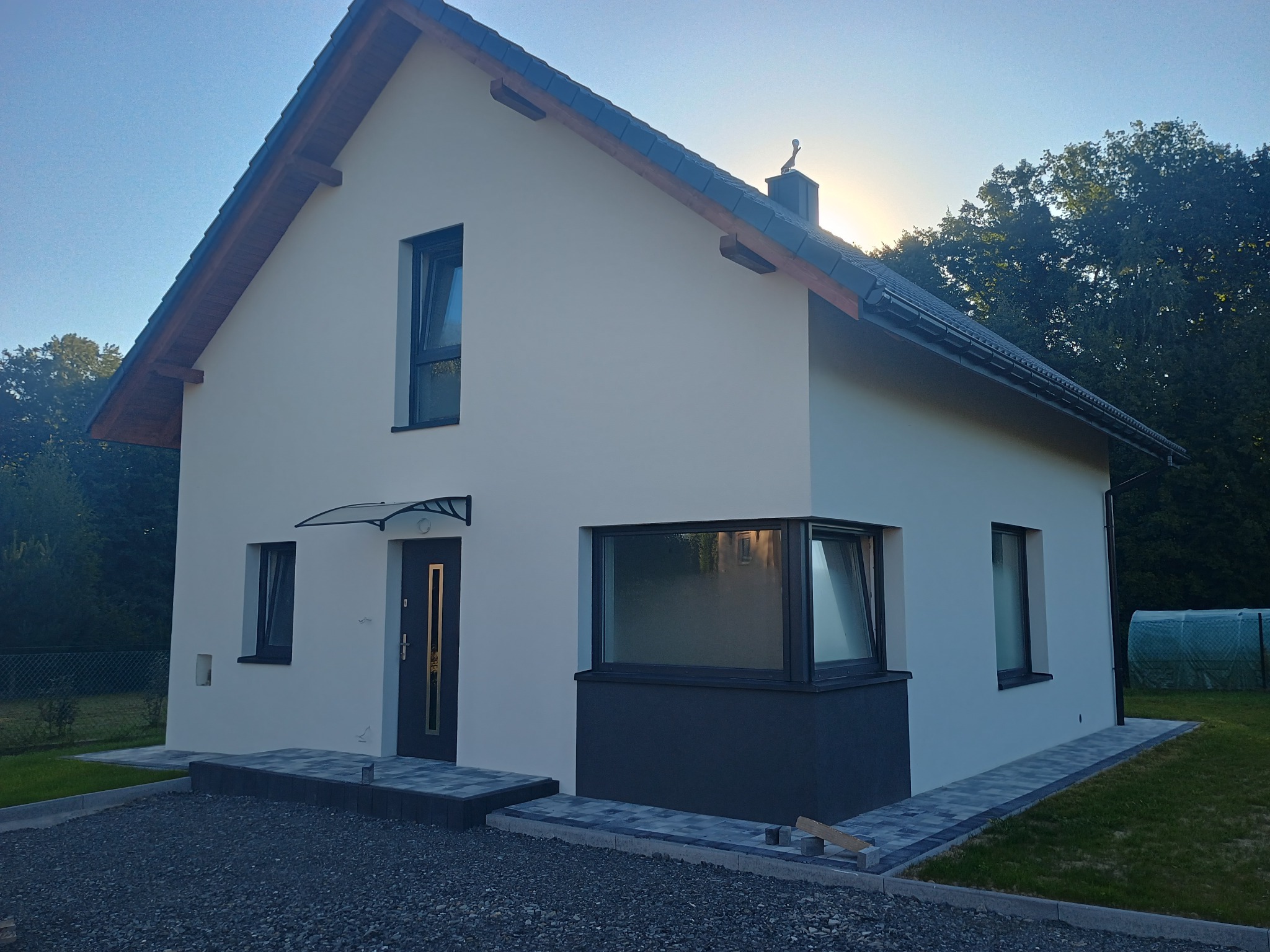 Dwukondygnacyjny dom z jasną elewacją, ciemną stolarką okienną i wejściem z zadaszeniem. Ułożona kostka brukowa wokół budynku. W tle zieleń drzew.