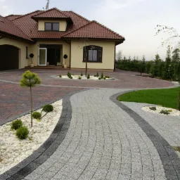 Elegancki podjazd i chodnik z kostki granitowej prowadzący do domu w Sosnowcu. Obrzeża z ciemnej kostki, jasna kostka na chodniku, czerwona na podjeździe.