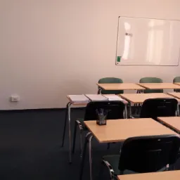 Pusta sala lekcyjna z biurkami, krzesłami, tablicą suchościeralną i stosem dokumentów na jednym z biurek.