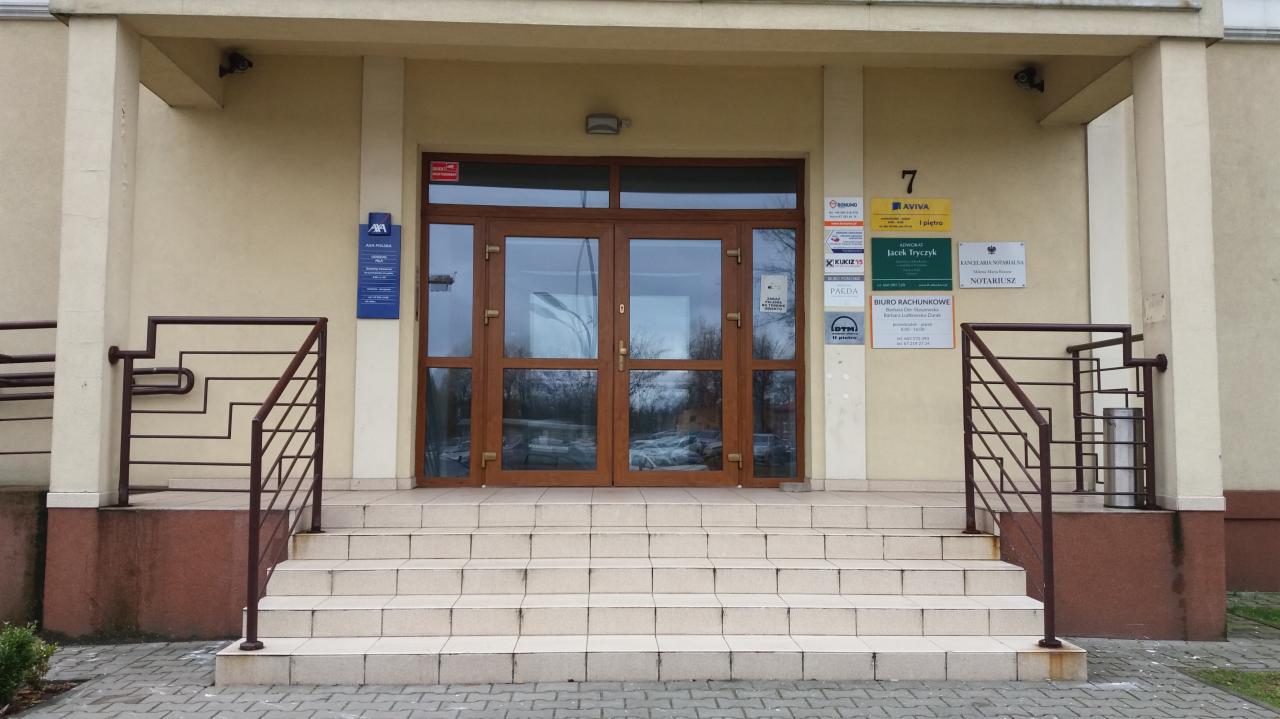 Wejście do budynku z tabliczkami informacyjnymi różnych firm i instytucji, w tym biura rachunkowego i szkoły jazdy, widok z dołu schodów.