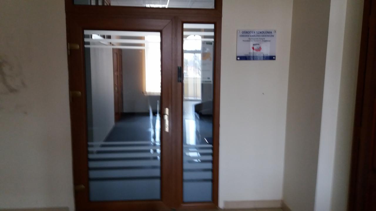 Wejście do placówki szkoleniowej z brązowymi drzwiami i tabliczką informacyjną na ścianie.