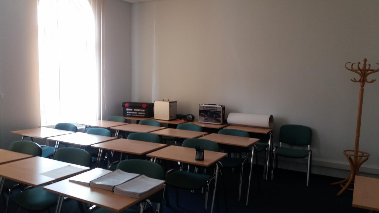 Sala wykładowa z rzędami stolików i krzeseł, na stolikach leżą notatniki, w tle widoczny wieszak na ubrania i skrzynki z wyposażeniem dydaktycznym.