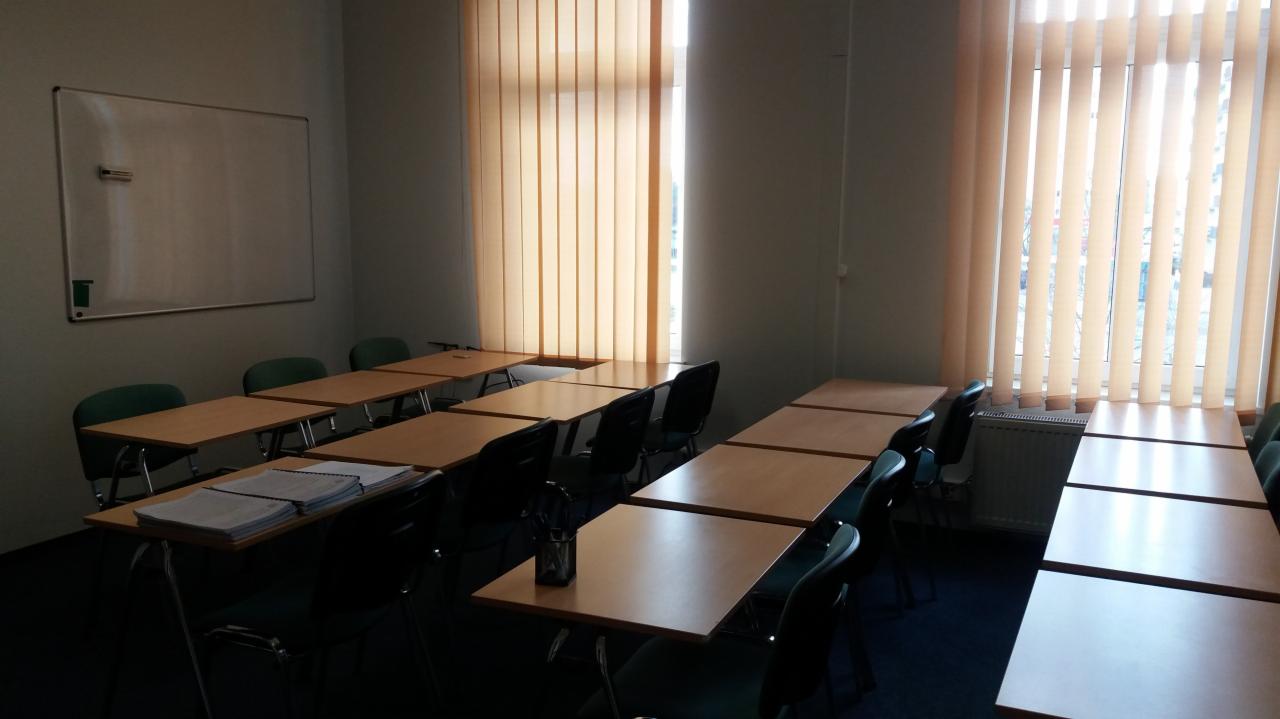Sala wykładowa z rzędami stolików i krzeseł, tablicą suchościeralną i pionowymi żaluzjami w oknach.