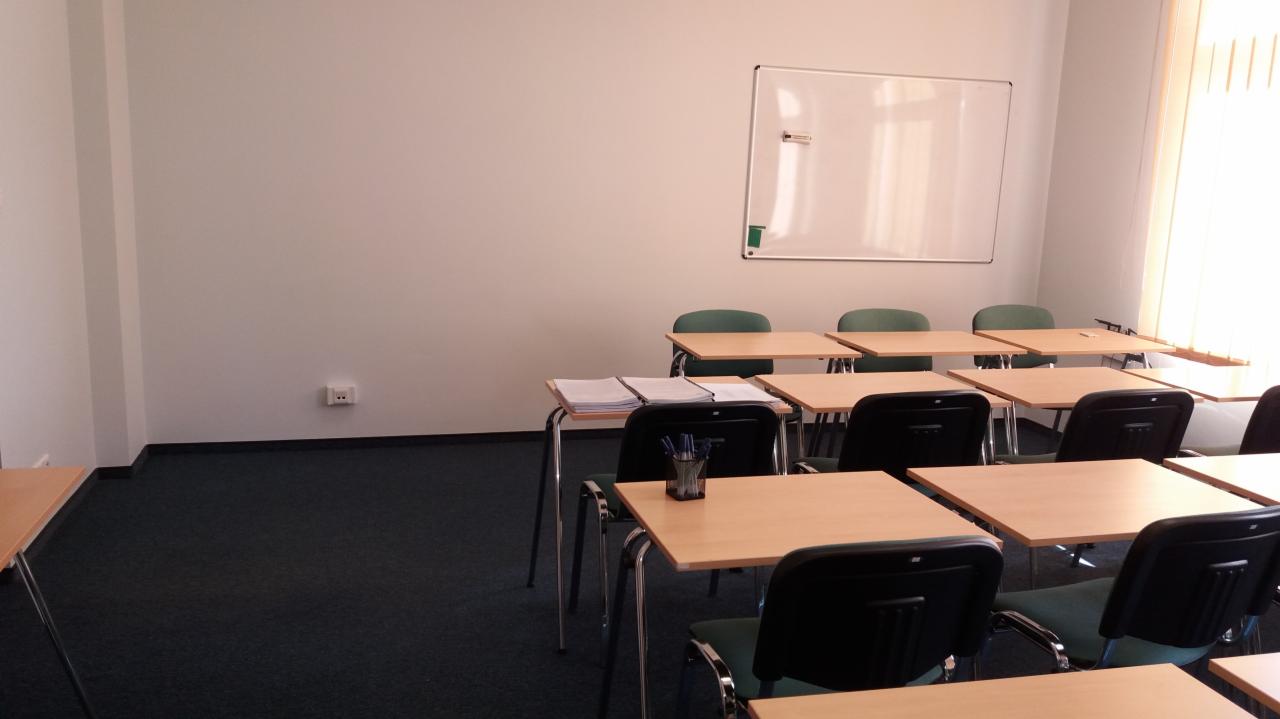 Pusta sala lekcyjna z biurkami, krzesłami, tablicą suchościeralną i stosem dokumentów na jednym z biurek.