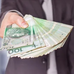 Dłoń w mankiecie marynarki trzymająca wachlarz zielonkawych banknotów 100 zł na rozmytym tle biurowym.