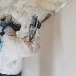 Pracownik w kombinezonie ochronnym aplikuje piankę izolacyjną na poddasze za pomocą pistoletu natryskowego, widoczne świeże krople pianki na ścianie.