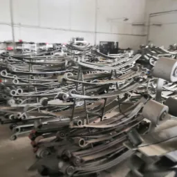 Stos metalowych resorów piórowych w magazynie warsztatu samochodowego, widoczny wózek widłowy w tle.