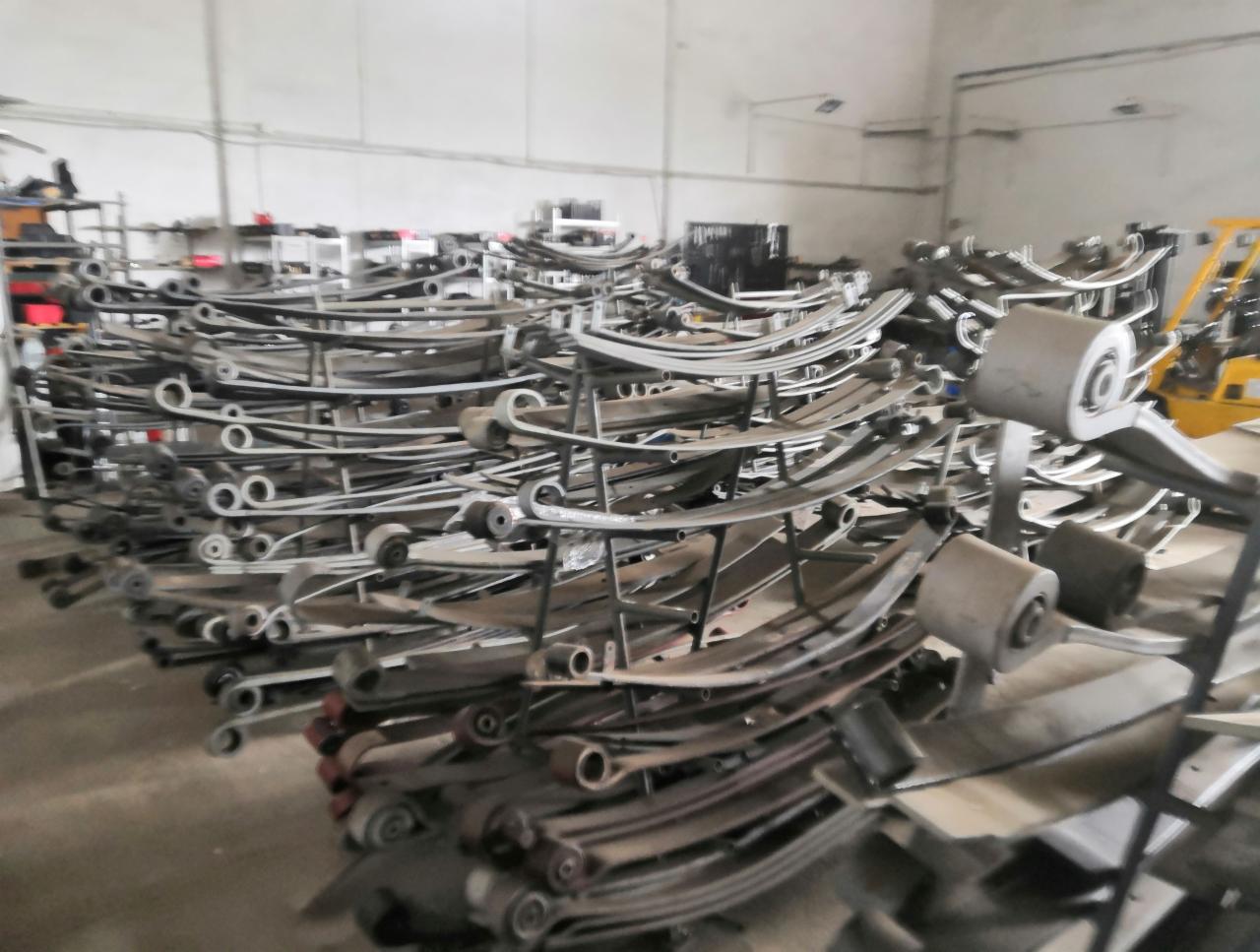 Stos metalowych resorów piórowych w magazynie warsztatu samochodowego, widoczny wózek widłowy w tle.