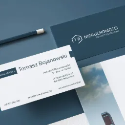 Wizytówki firmy TB Nieruchomości z logo, danymi kontaktowymi i adresem, ułożone na ciemnoniebieskim tle obok ołówka.