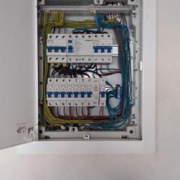 Grzegorz Szymczak - Otwarta skrzynka bezpiecznikowa z widocznymi bezpiecznikami, przewodami i listwami zaciskowymi, zamontowana na białej ścianie.