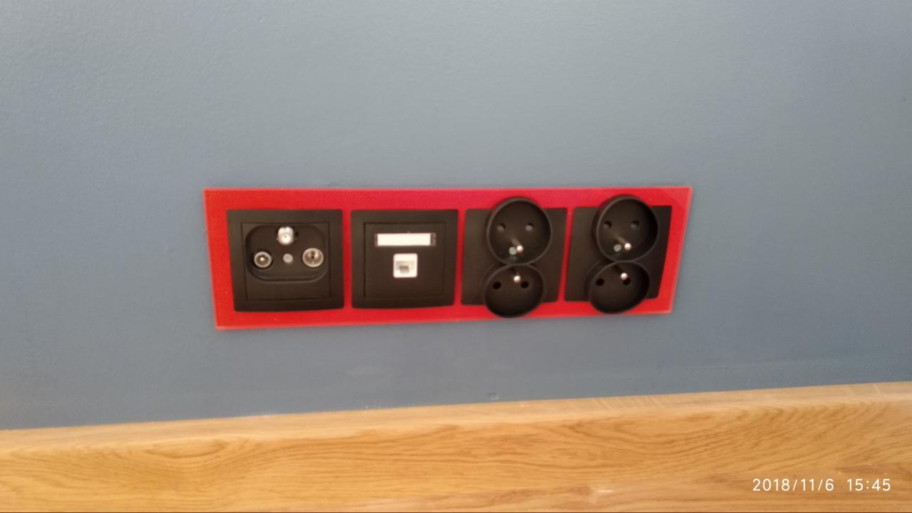 Rząd czarnych gniazdek i wtyczek, w czerwonych ramkach, zamontowanych na szarej ścianie nad drewnianą listwą przypodłogową.