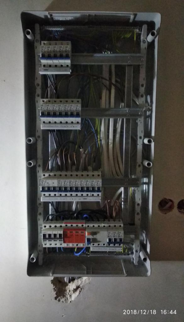 Rozdzielnica elektryczna w trakcie montażu, widoczne przewody, bezpieczniki i listwy zaciskowe, osadzona w szarej obudowie w ścianie z widocznymi ubytkami tynku.