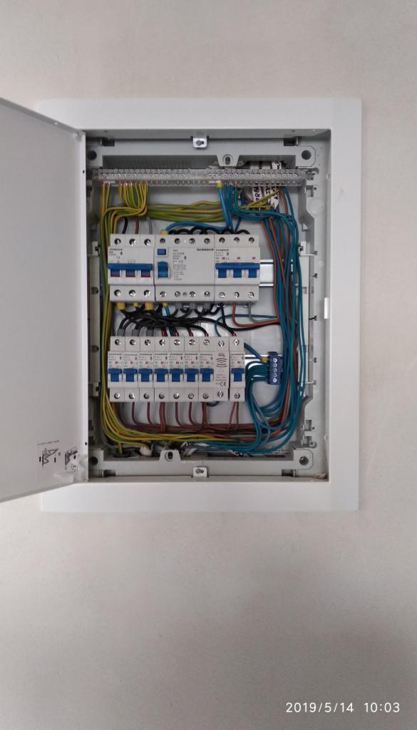 Otwarta skrzynka bezpiecznikowa z widocznymi bezpiecznikami, przewodami i listwami zaciskowymi, zamontowana na białej ścianie.