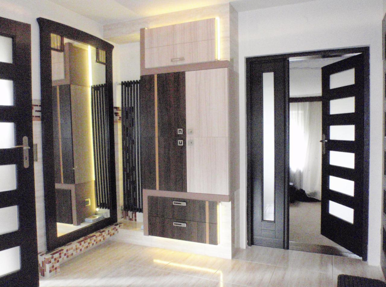 Nowoczesna aranżacja łazienki z zabudową meblową w odcieniach beżu i brązu z podświetleniem LED, widoczne drzwi z przeszkleniami i dekoracyjne mozaiki przy podłodze.