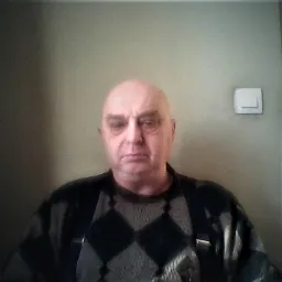 Portret mężczyzny w swetrze w romby na tle ściany z włącznikiem światła.